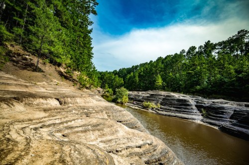 A serene river flows through layered rock formations, surrounded by lush green trees under a blue sky.
