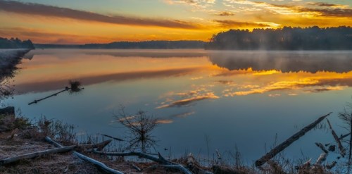 A serene lake at sunrise, with colorful reflections and mist rising from the water, surrounded by trees and logs.