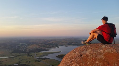 A person sits on a rock at sunset, overlooking a scenic landscape with hills and a lake.