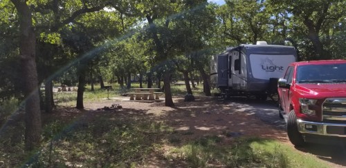 A red truck parked next to a camper in a wooded campsite with picnic tables and greenery surrounding.