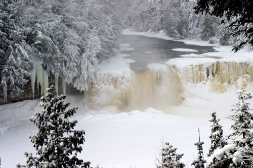 A frozen waterfall surrounded by snow-covered trees and mist rising from the water, creating a serene winter scene.