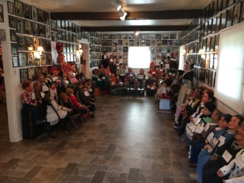 A room filled with various dolls and mannequins seated in rows, surrounded by framed photographs on the walls.
