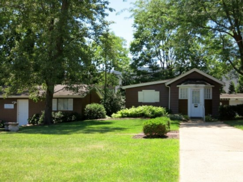 Two houses with green lawns and trees, one brick and one brown, set in a sunny residential area.