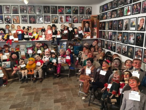 A room filled with various ventriloquist dummies and puppets, surrounded by framed photos on the walls.