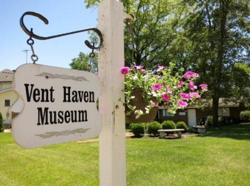 Sign for Vent Haven Museum with hanging flowers in a green, sunny yard.