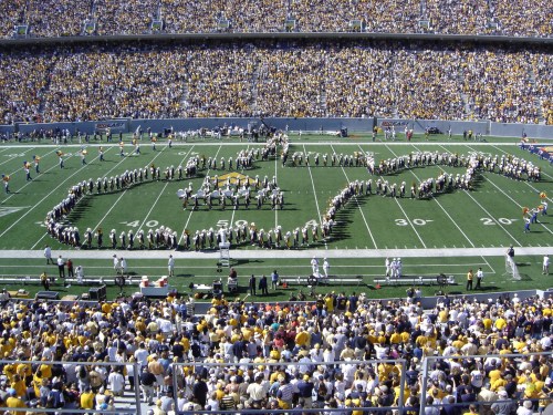 A large crowd at a football stadium watches a marching band form a musical note on the field.