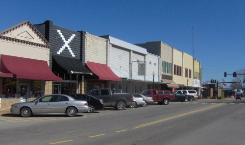 A small town street lined with shops, featuring parked cars and colorful awnings under a clear blue sky.