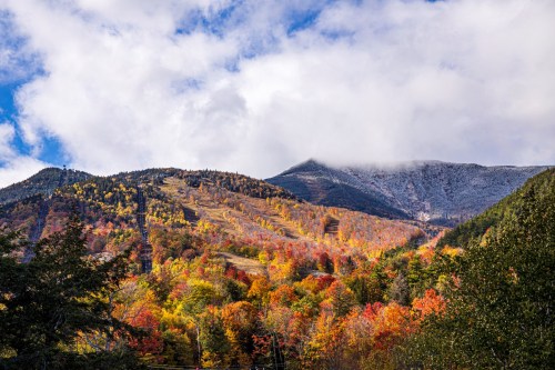 Vibrant autumn foliage covers mountains under a partly cloudy sky, showcasing a mix of orange, yellow, and green trees.