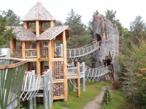 A wooden play structure with a tower and rope bridge, surrounded by trees and greenery.