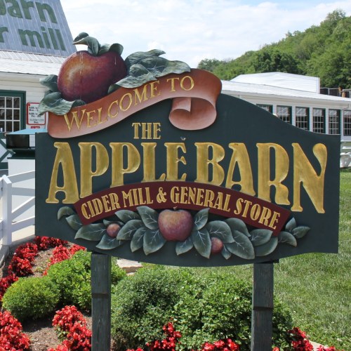 Sign welcoming visitors to The Apple Barn Cider Mill & General Store, surrounded by colorful flowers and greenery.