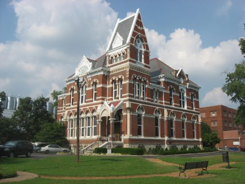 Historic brick building with ornate architecture, surrounded by greenery and a park, under a partly cloudy sky.