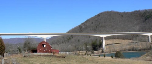 A red barn sits in a field beneath a large, elevated bridge with mountains in the background and a pond nearby.
