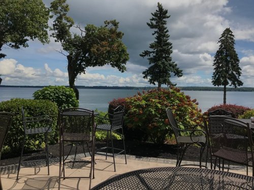 A serene lakeside view with trees, colorful shrubs, and empty chairs on a patio under a partly cloudy sky.