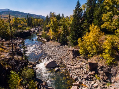 A serene river flows through a rocky landscape, surrounded by trees with autumn foliage under a clear blue sky.