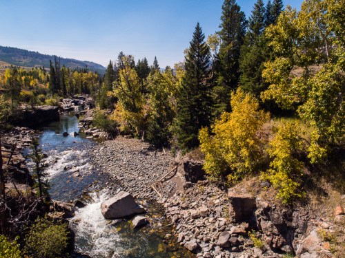 A serene river flows through a rocky landscape, surrounded by trees with vibrant autumn foliage under a clear blue sky.