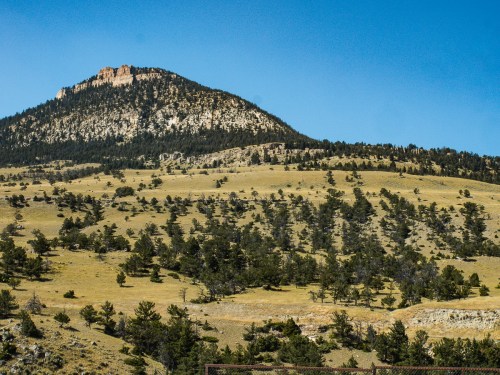 A scenic view of a mountain with a rocky peak, surrounded by rolling hills and scattered trees under a clear blue sky.