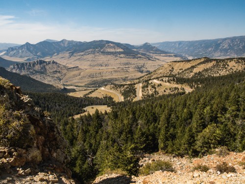 A panoramic view of rugged mountains and valleys, with dense forests and winding dirt roads under a clear blue sky.