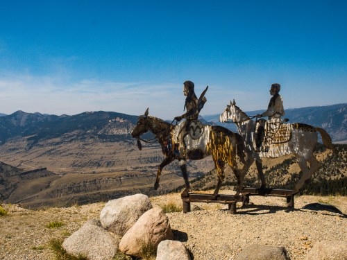 Metal sculptures of two riders on horseback overlook a vast mountainous landscape under a clear blue sky.