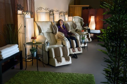 A woman relaxes in a massage chair surrounded by soft lighting and greenery in a tranquil spa setting.