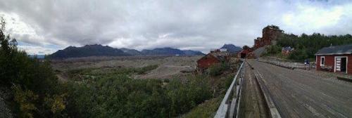 Panoramic view of a rugged landscape with mountains, a wooden path, and remnants of red buildings among greenery.