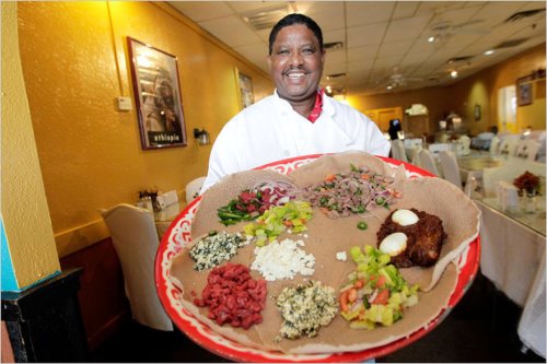A chef proudly holds a large platter of colorful Ethiopian dishes, featuring injera and various toppings, in a restaurant.