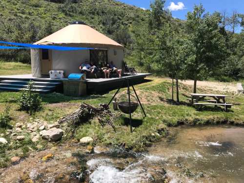 A yurt by a stream, with people sitting on the deck, surrounded by greenery and a picnic table nearby.