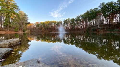 A serene lake surrounded by trees, reflecting the sky and a fountain in the distance, with rocks in the foreground.