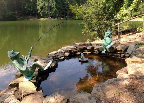 Two bronze frog sculptures by a pond, one fishing and the other watching, surrounded by greenery and rocks.