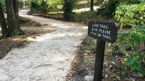 A wooden sign on a path reads "Lake Trail 5.1 mi Please Stay on Trail," surrounded by trees and greenery.