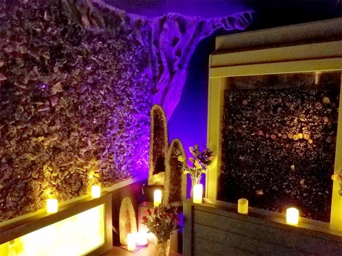 A serene spa room with crystal walls, soft purple lighting, candles, and decorative plants for a calming atmosphere.