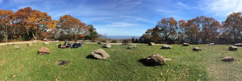 A panoramic view of a grassy area with large rocks, surrounded by autumn trees and a clear blue sky.