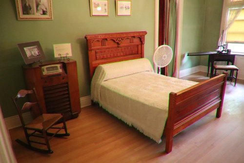 A vintage bedroom featuring a wooden bed, rocking chair, fan, and a small desk against a green wall.