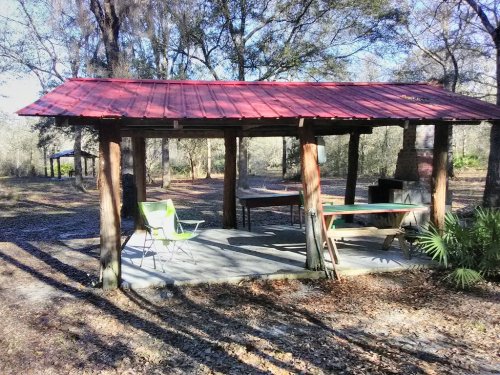 A rustic outdoor shelter with a red roof, wooden posts, and a green chair, surrounded by trees and fallen leaves.