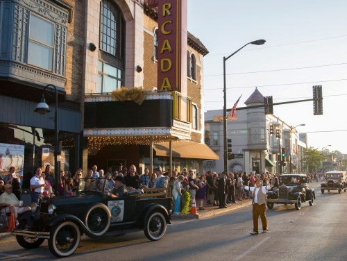 A vintage car parade in front of a theater, with a crowd watching and buildings lining the street in the background.