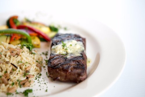 A grilled steak topped with butter, served with rice and colorful sautéed vegetables on a white plate.