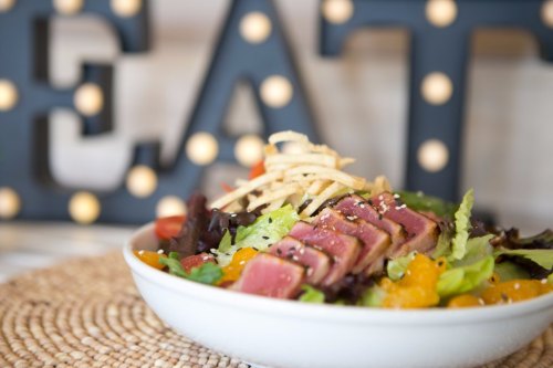 A vibrant salad with seared tuna, mixed greens, and crispy toppings, set against a decorative "EAT" sign.