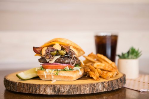 A juicy burger with bacon, cheese, and toppings on a wooden platter, served with fries and a drink.