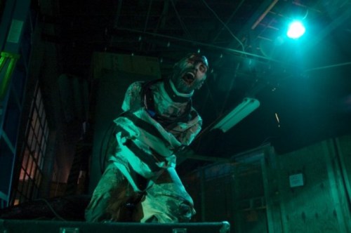 A man in a straitjacket screams in a dimly lit, industrial setting, with eerie green lighting.