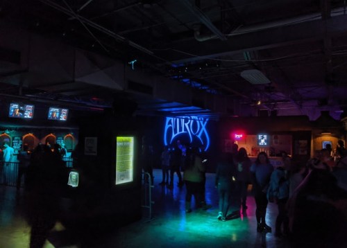 Dimly lit venue with a neon "AURORA" sign, people mingling, and various screens displaying information.