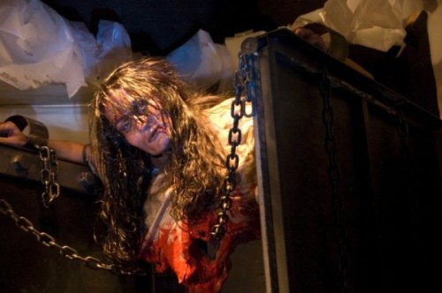 A horror scene featuring a figure with long hair, appearing injured and chained in a dark, eerie environment.