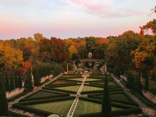 A beautifully landscaped garden with geometric hedges, surrounded by vibrant autumn trees and a serene pond at sunset.