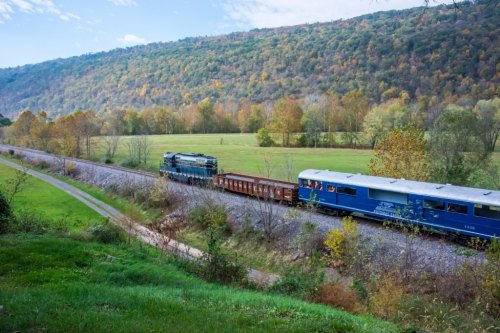 A train travels along a scenic route with rolling hills and autumn foliage in the background.