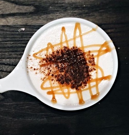 A small white dish with a creamy dessert topped with caramel drizzle and a sprinkle of cocoa powder.