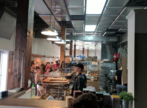 A busy bakery interior with customers in line and baked goods displayed on the counter. Bright, industrial-style decor.