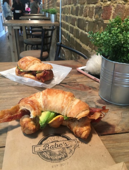A croissant sandwich with bacon and avocado on a wooden table, with another sandwich and greenery in the background.