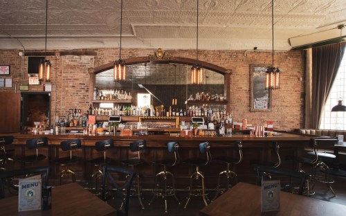A cozy bar with a wooden counter, high stools, and a mirror backdrop, featuring brick walls and warm lighting.
