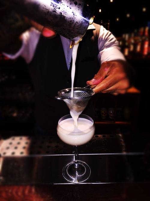 A bartender pours a cocktail through a strainer into a glass, with a blurred bar background.