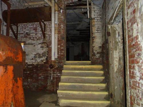 Dimly lit basement with brick walls, a staircase leading up, and an orange industrial object on the left.