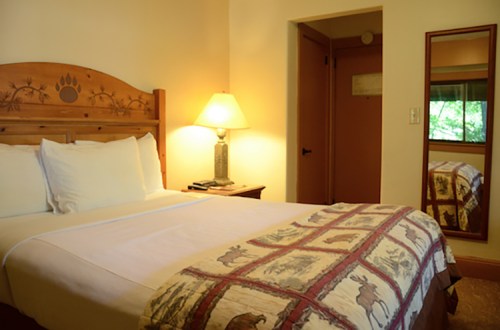 Cozy hotel room with a wooden bed, decorative quilt, lamp, and a mirror reflecting the entrance.