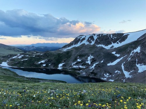 A serene mountain landscape with a lake, surrounded by snow-capped peaks and colorful wildflowers in the foreground.